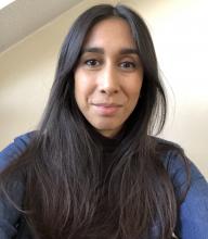 A portrait of a light-skinned south asian woman with long straight brown-black hair, wearing a blue shirt against a plain light background 